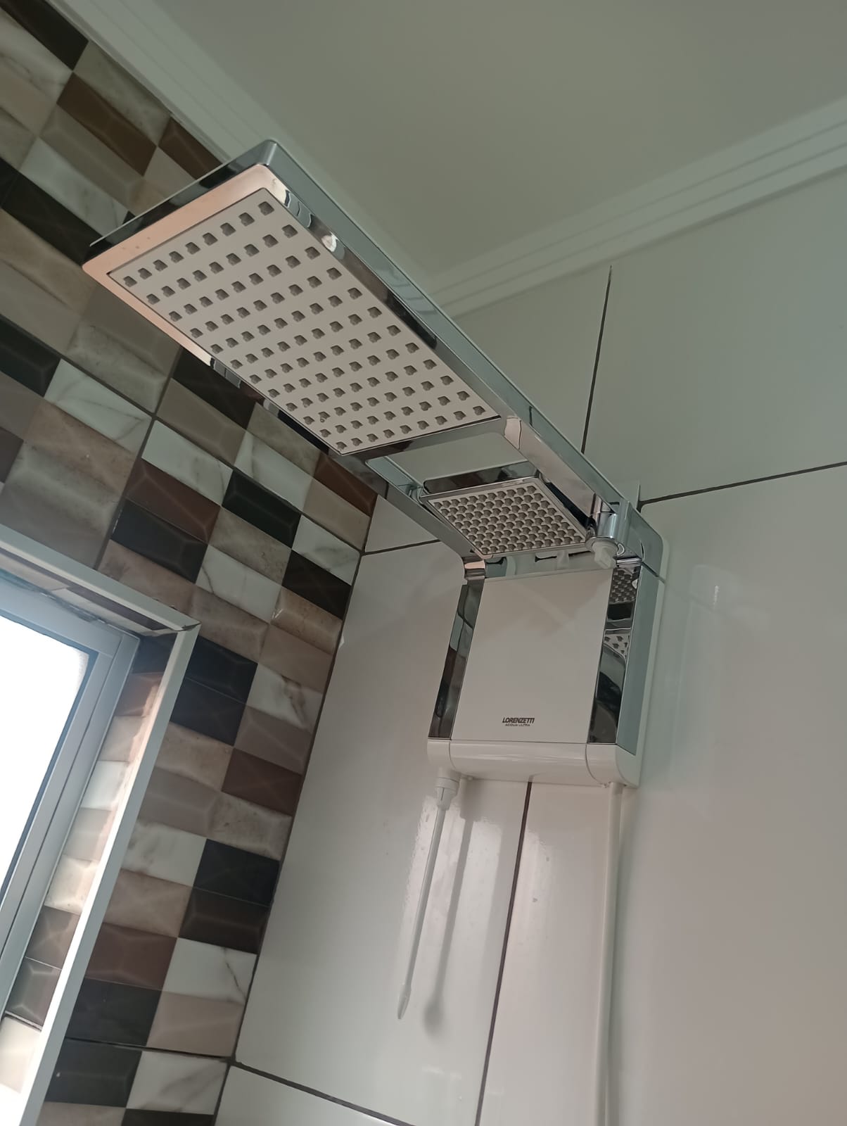 ducha quadrada Lorenzetti cromada instalada em banheiro com azulejos em tons de mármore e janela ao lado