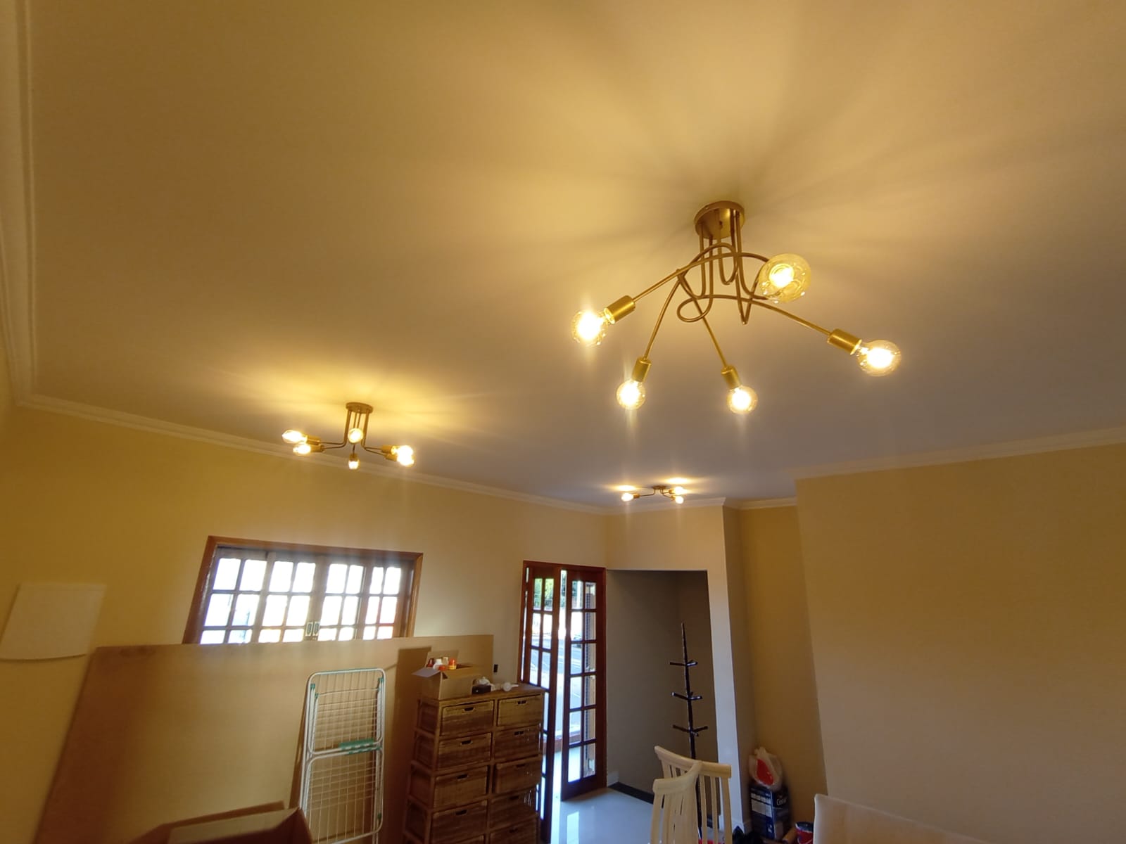 sala com três luminárias douradas de teto acesas, com hastes curvas e lâmpadas de filamento, paredes bege e po